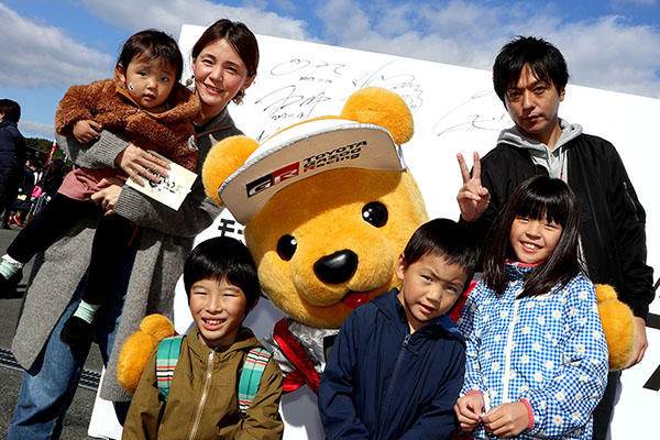 トヨタ くま吉 グレーのニットコートを着た女性に抱き上げられる女の子と、くま吉の前に3人で並ぶ子たちに黒い上着を着た男性の集合写真@ モースポフェス in 九州